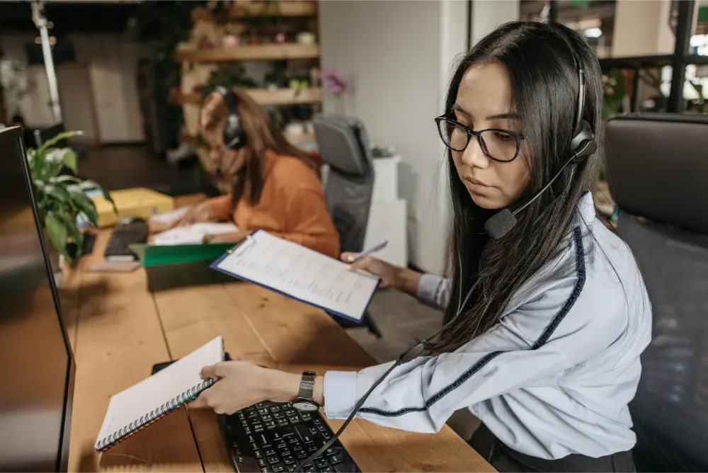 Two women wearing headsets work at desks in an MSP office. The woman in the foreground looks at a clipboard and types on a keyboard, while the woman in the background writes on paper. Both are focused on their tasks.