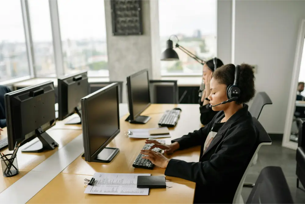 Two women wearing headsets work at desks with computers in a bright, modern MSP office. One types on a keyboard while the other listens attentively. Office supplies and documents are neatly arranged on the desk.