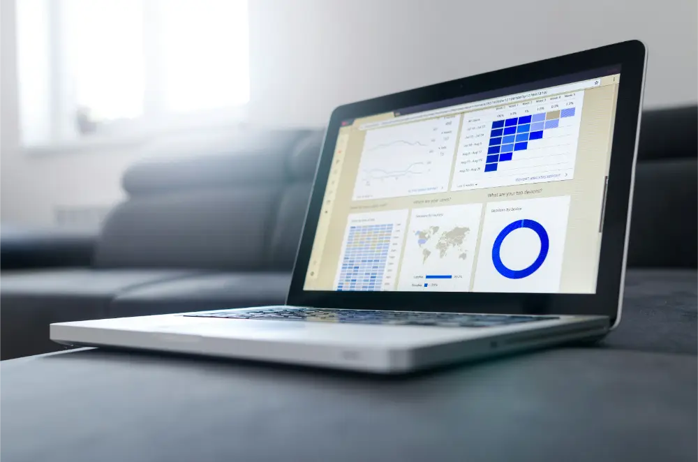 A laptop on a gray sofa displays various data visualizations, including MSP performance graphs, charts, a world map, and a blue pie chart. The background is softly lit with natural light from a window.