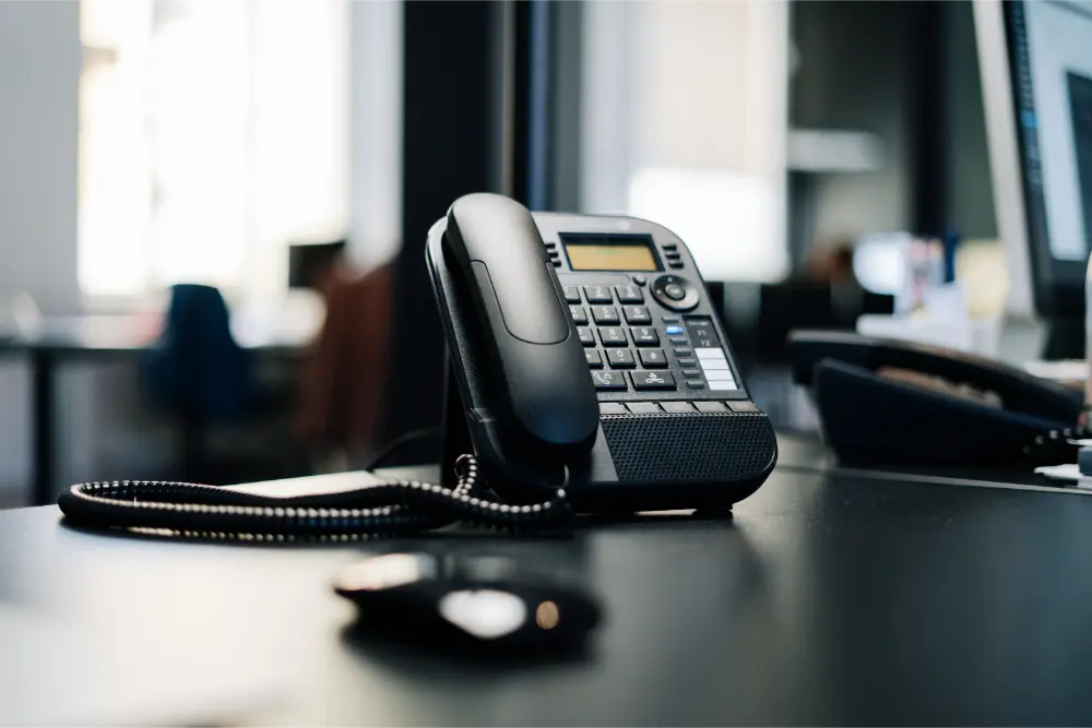 A black office desk phone with a digital display sits on a desk, its corded handset resting in place—ideal for any MSP environment. The background shows a blurred modern office setting with computers and furniture.