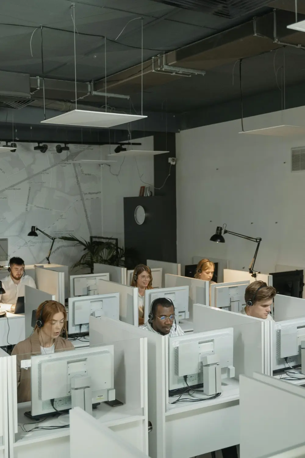 Several people wearing headsets work at computers in a modern MSP office with individual cubicles, focused on their screens. The workspace is well-lit and organized, with lamps and overhead lighting.