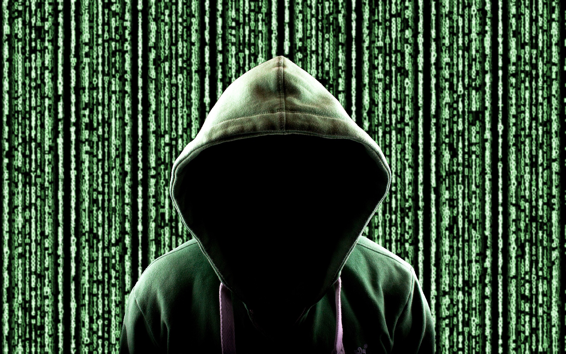 A person in a green hoodie with the hood up, obscuring their face in darkness, stands in front of a background featuring vertical green lines of digital code.