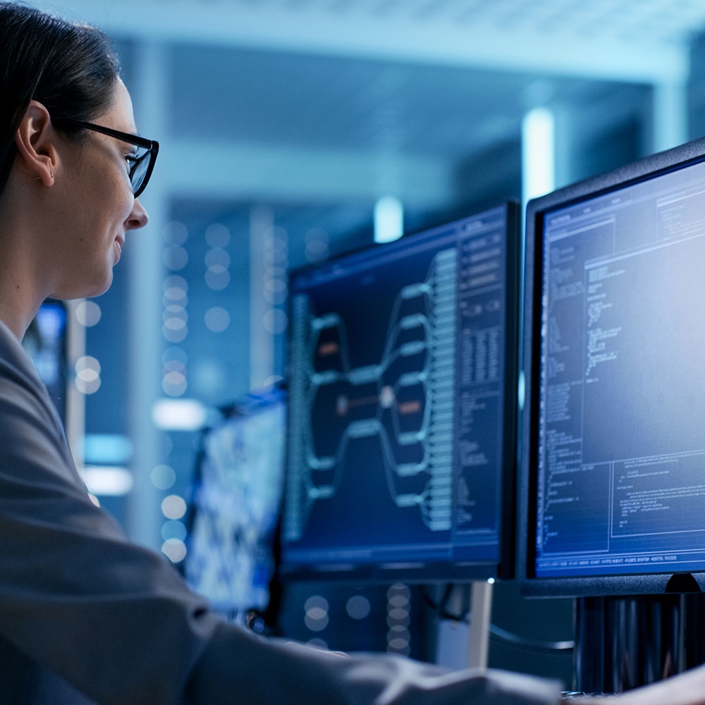 A person wearing glasses and a business suit works at a desk with multiple computer screens displaying complex data and diagrams.