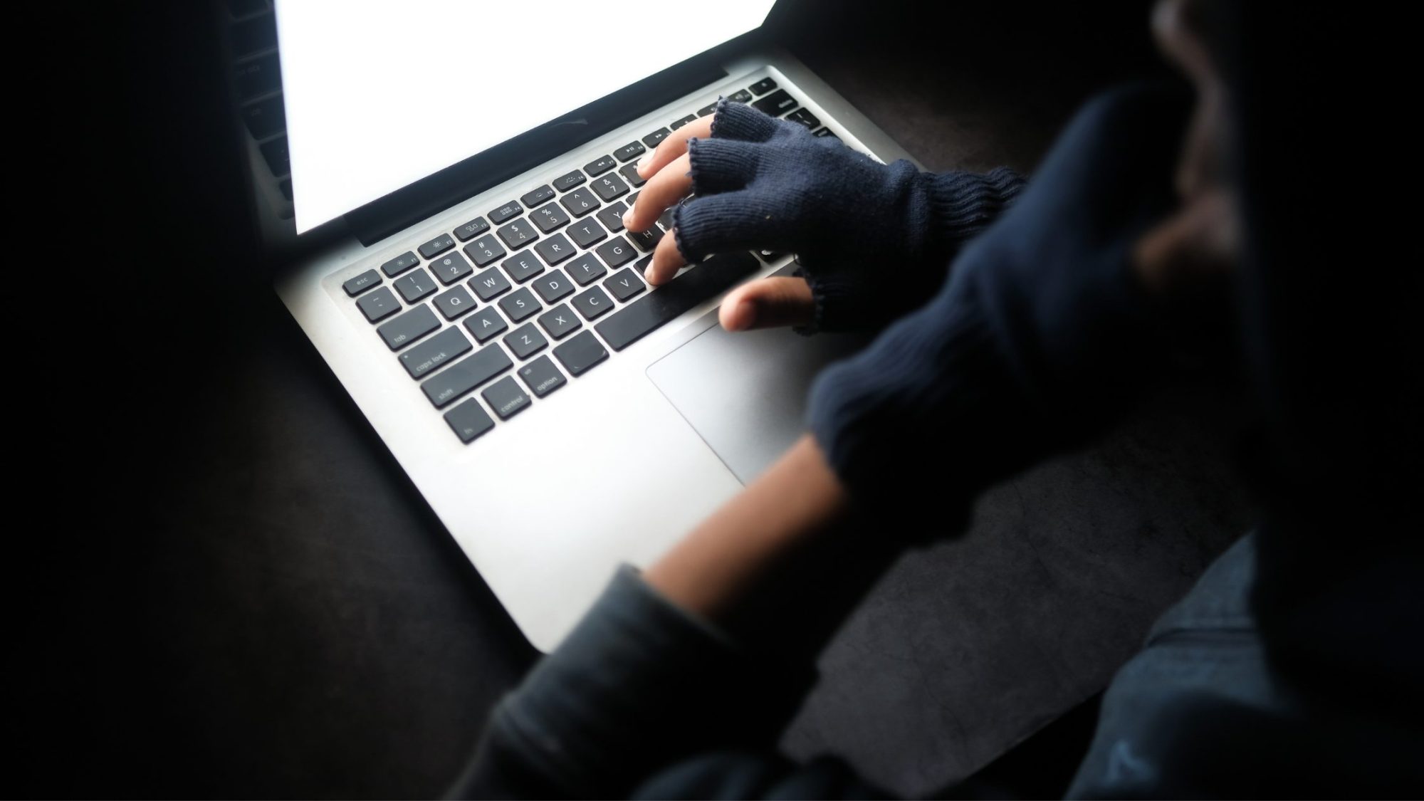 A person wearing fingerless gloves is typing on a laptop in a dimly lit environment, clearly immersed in cybersecurity operations.