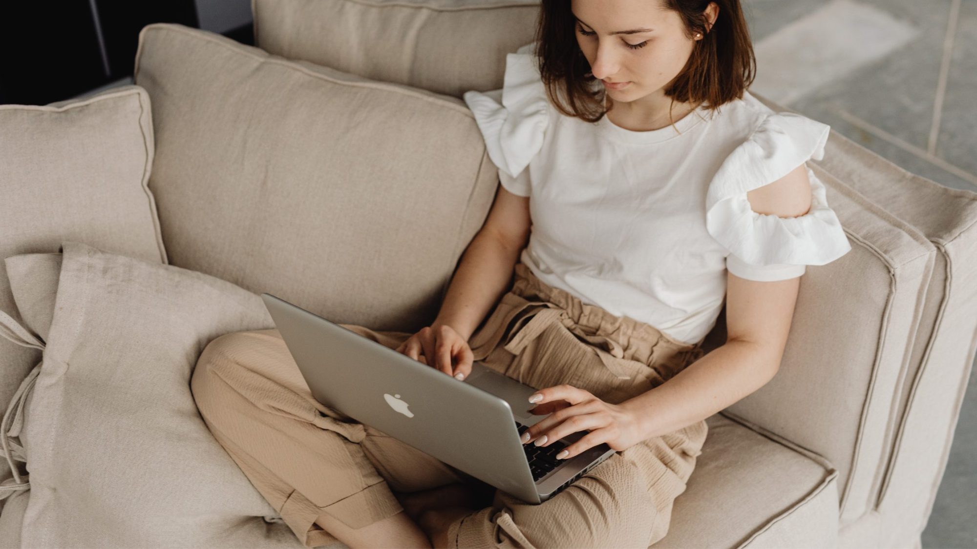 A woman sits on a beige couch with her legs crossed, working on a silver laptop. 