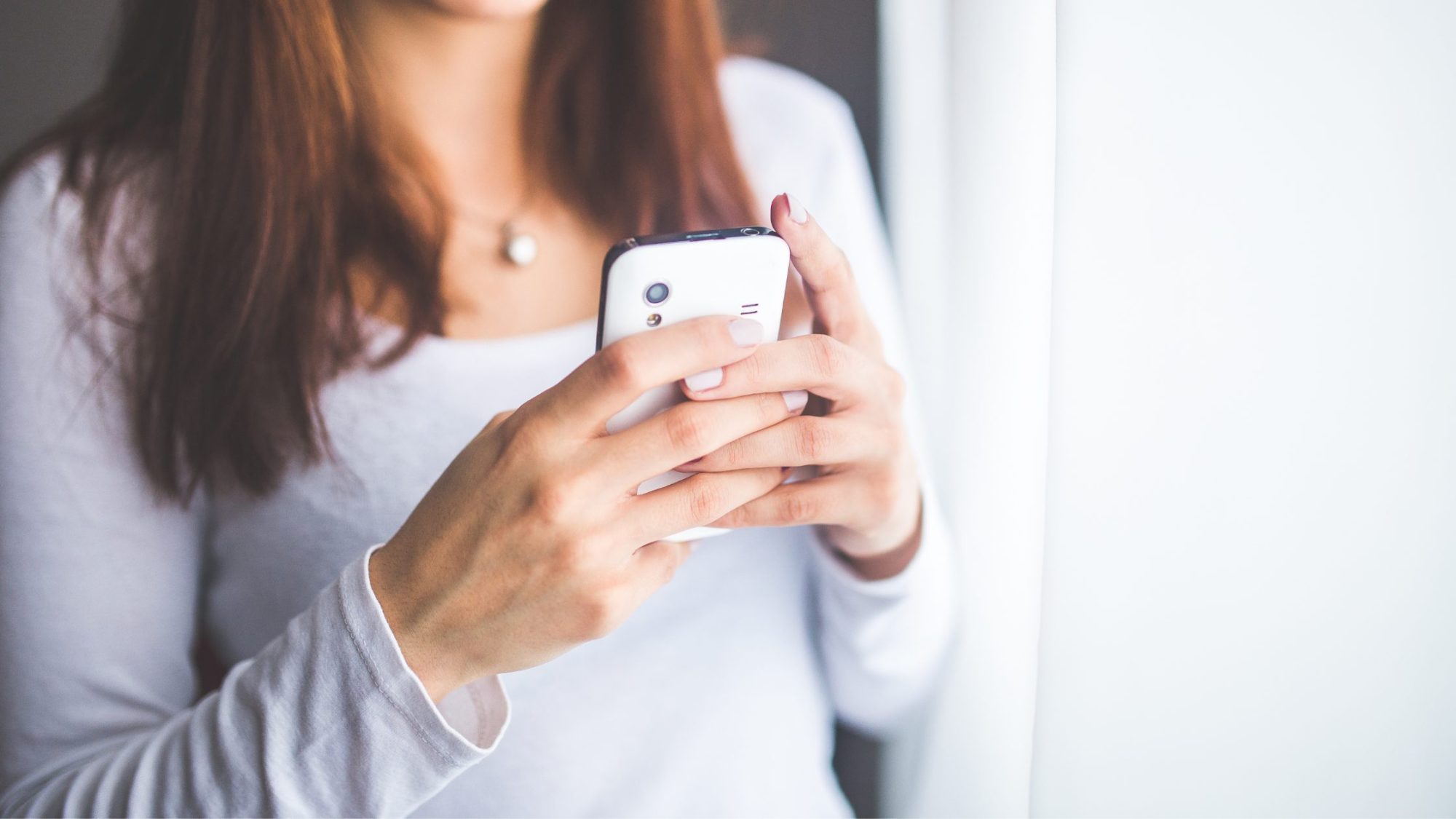 A woman with long brown hair, wearing a white long-sleeve top, holds and looks at a smartphone.