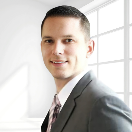 A man in a gray suit with a white shirt and patterned tie is standing in front of large windows emitting natural light. He has short dark hair and is smiling slightly while looking into the camera. The background is minimalistic and white.