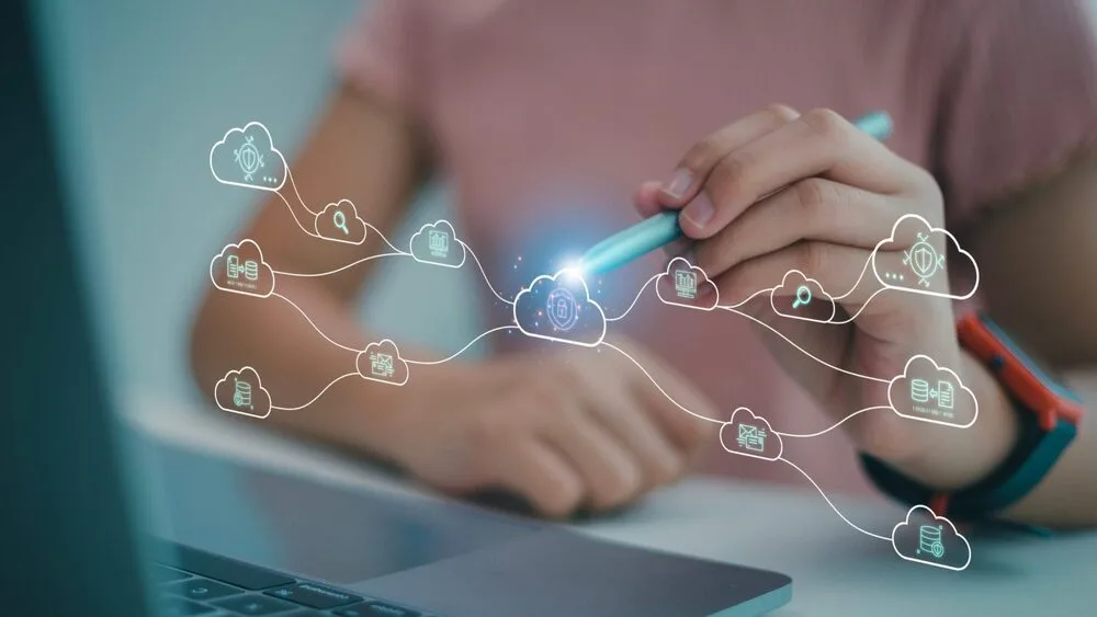 Person using a stylus to interact with a network of cloud icons displayed as holograms above a laptop keyboard.