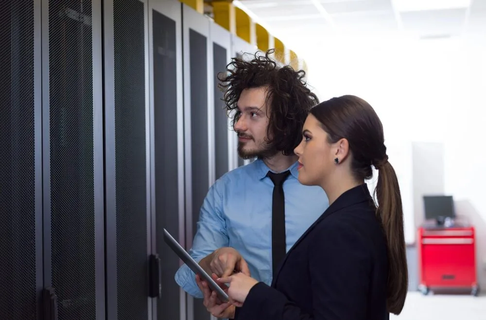 Two people in business attire, a man with curly hair pointing at something while holding a tablet, and a woman with a ponytail looking on, are standing in a server room with racks of servers.