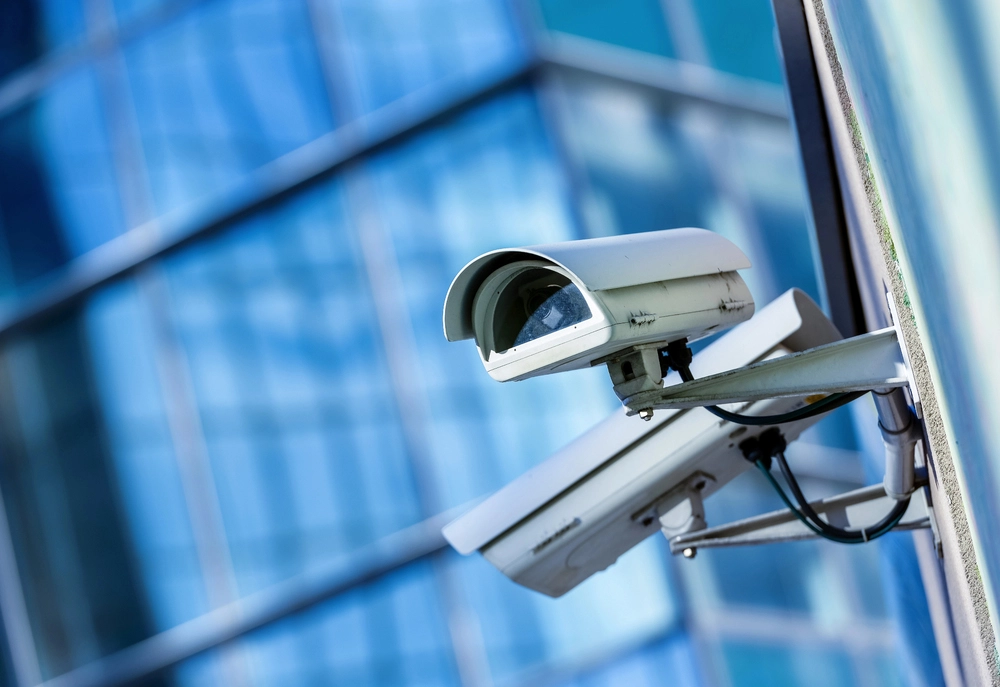 Close-up of a white security camera mounted on a building exterior. The background shows a glass facade reflecting blue and green hues, suggesting an urban setting with modern architecture.