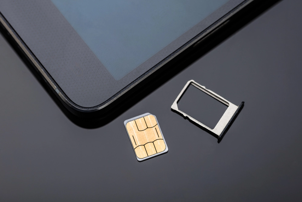 A close-up image of a SIM card and SIM card tray placed on a shiny black surface next to a tablet. The tablet screen is partially visible, with the SIM card and tray positioned beside it.