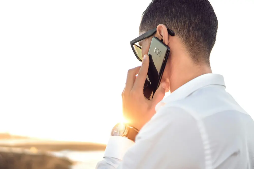 A person wearing glasses and a white shirt is outdoors during sunset, holding a smartphone to their ear with their left hand. The sunlight is hitting the phone, creating a bright reflection. The person appears to be engaged in a phone conversation.