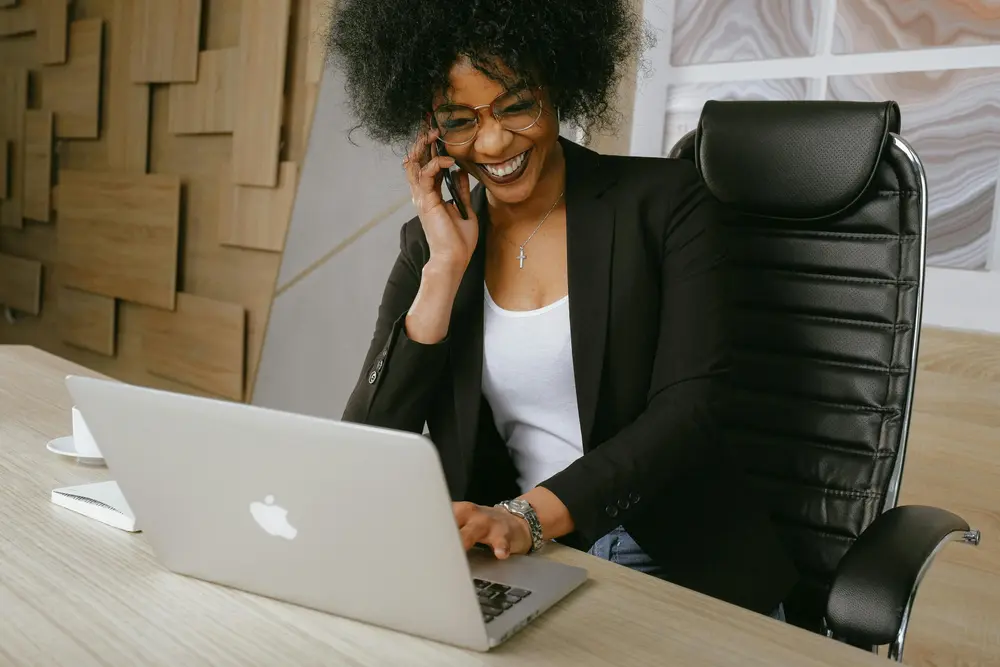 A person with curly hair, wearing glasses and a black blazer, smiles while talking on a cellphone. They are seated at a desk in an office setting, typing on a silver laptop. A coffee cup and saucer are on the desk.