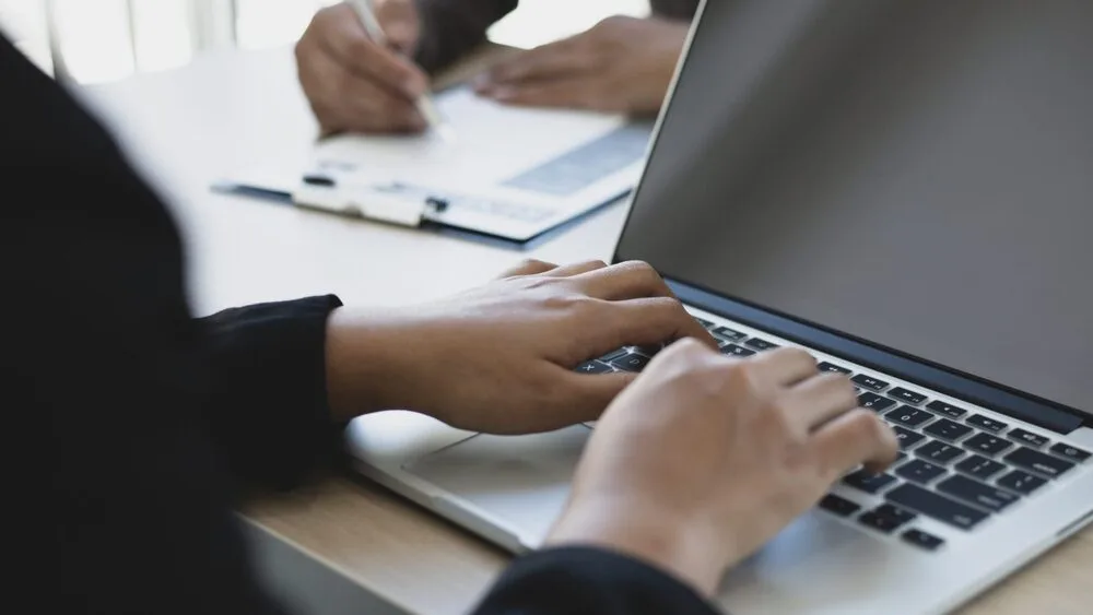 Close-up of hands typing on a laptop keyboard. In the background, a person is writing on a clipboard.