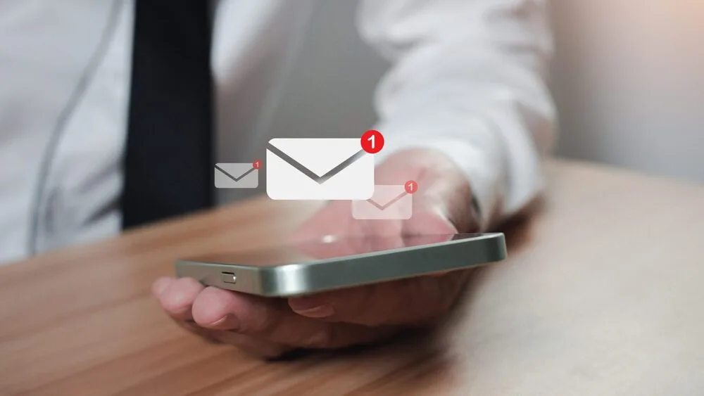 A person in a white shirt holding a smartphone screen up, displaying three email icons with notification badges. 