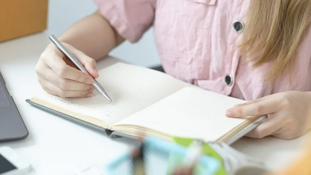 A person wearing a pink button-up shirt is writing in a notebook with a pen. The notebook is open on a white desk, and a laptop and some objects are partially visible in the foreground.