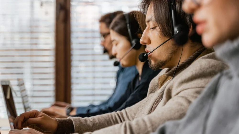 A row of people sitting at desks, each wearing headsets and working on laptops.