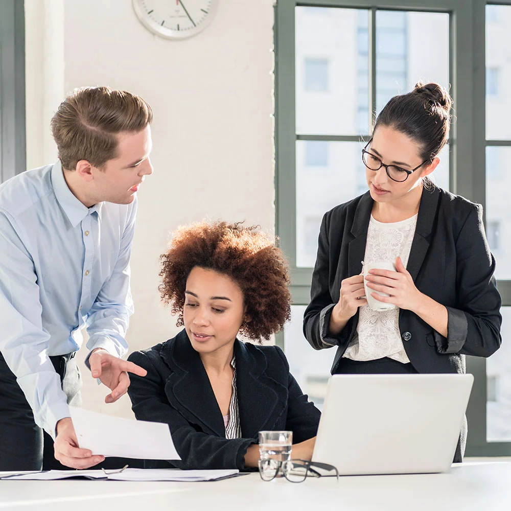 Three professionals in a bright office setting; a man pointing at a paper, a woman with curly hair looking at it, and another woman holding a notebook, standing by a laptop on the desk.