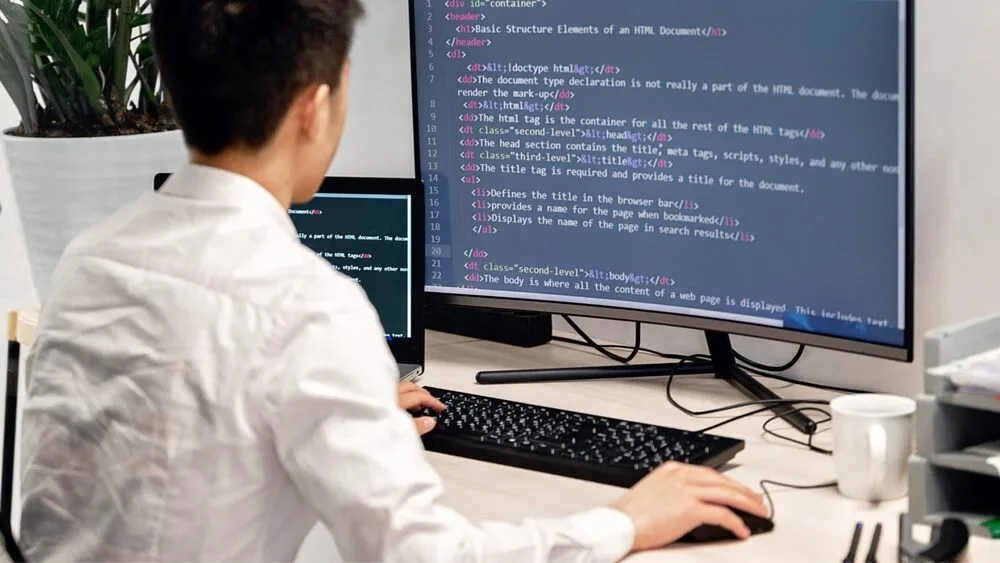 A person in a white shirt is sitting at a desk working on a computer with HTML code displayed on a large monitor.