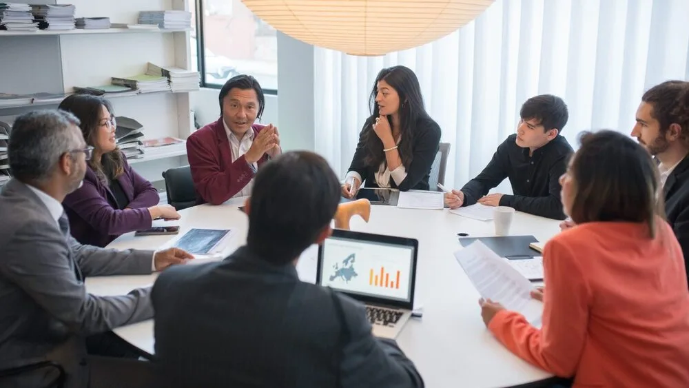 A diverse group of people sit around a round table in an office, engaged in a meeting.