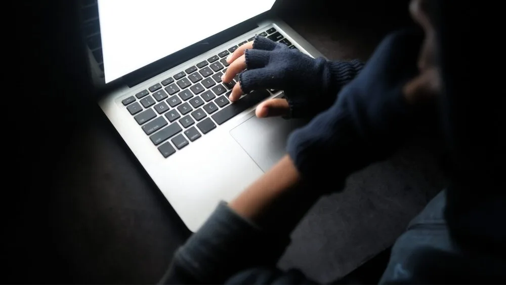 A person wearing fingerless gloves types on a laptop in a dark room. The screen emits a bright glow, illuminating their hands and casting shadows on the keyboard.