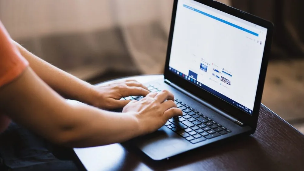 Person typing on a laptop resting on a wooden table, with a web browser open on the screen displaying a minimalistic website layout. 