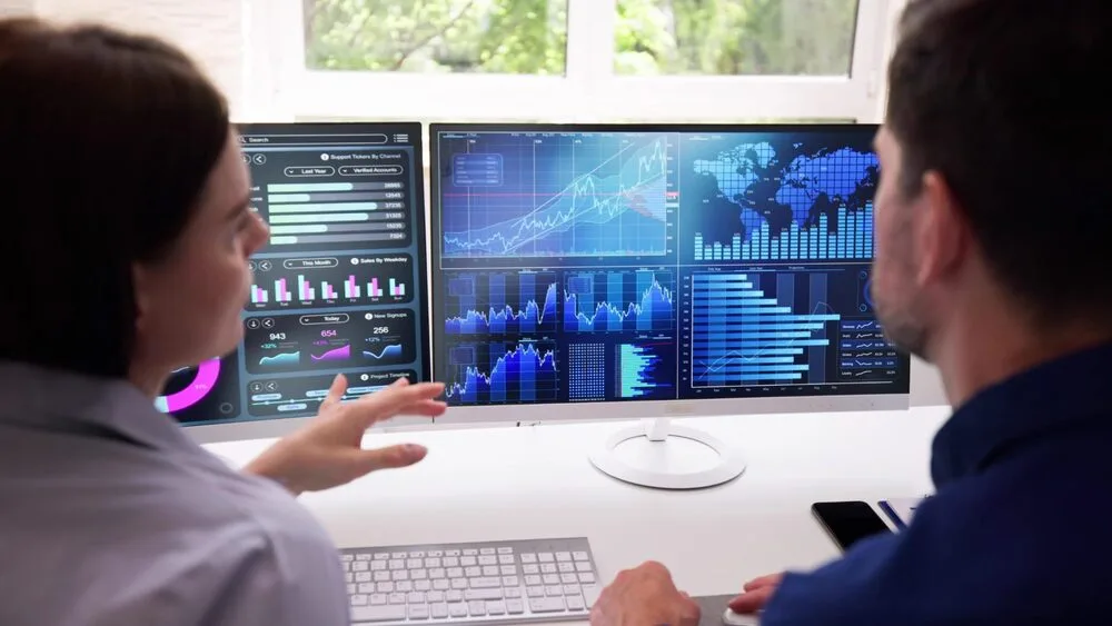 Two people are sitting at a desk with dual monitors displaying various data visualizations, including graphs and charts. 