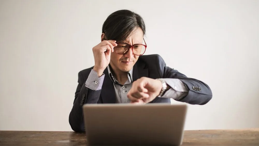 A person in a suit and glasses sits at a table, looking intently at their watch while working on a laptop. 