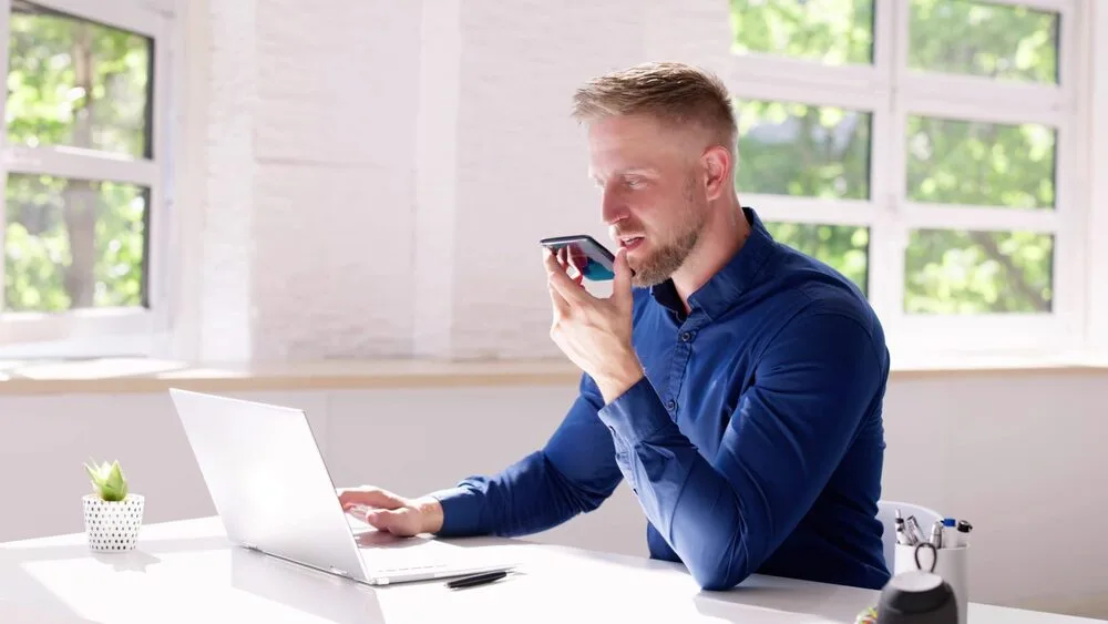 A man in a blue shirt sits at a white desk, using a laptop and speaking into his smartphone. 