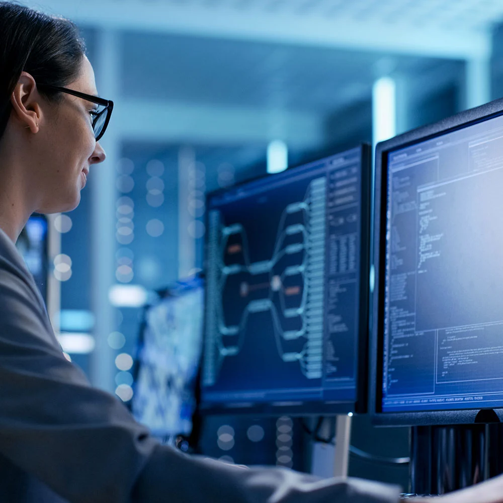 A person wearing glasses works at a computer desk with multiple screens displaying complex data and code in a dimly lit room. The background shows additional computer equipment, suggesting a high-tech or cybersecurity environment.