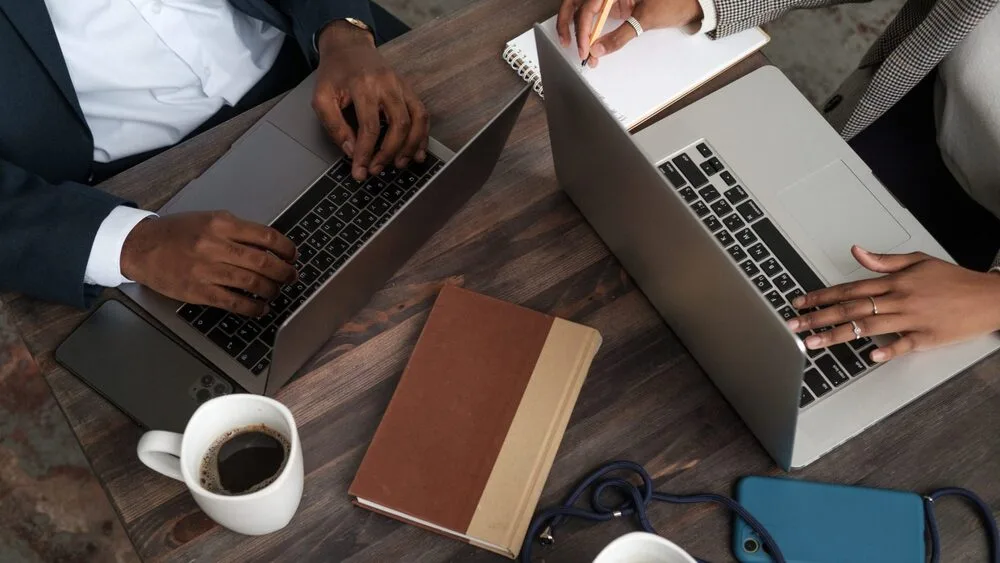 Two people are working at a wooden table with laptops open.