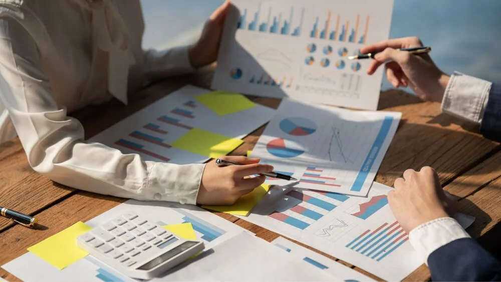 Two people sitting at a wooden table analyzing printed charts and graphs.