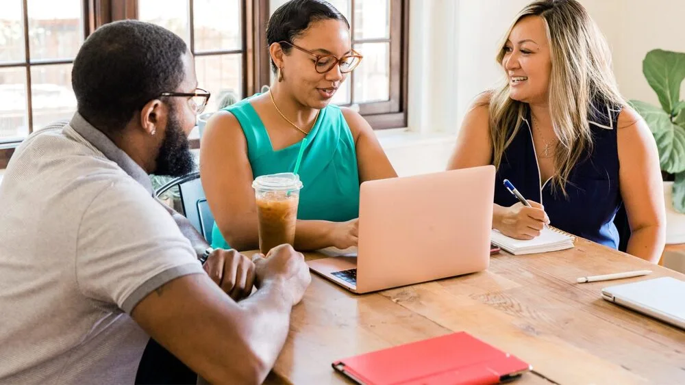 Three people sit around a wooden table in an office. One woman uses a laptop while holding an iced coffee. Another woman takes notes. A man listens attentively. Notebooks lie on the table. They appear engaged in a discussion.