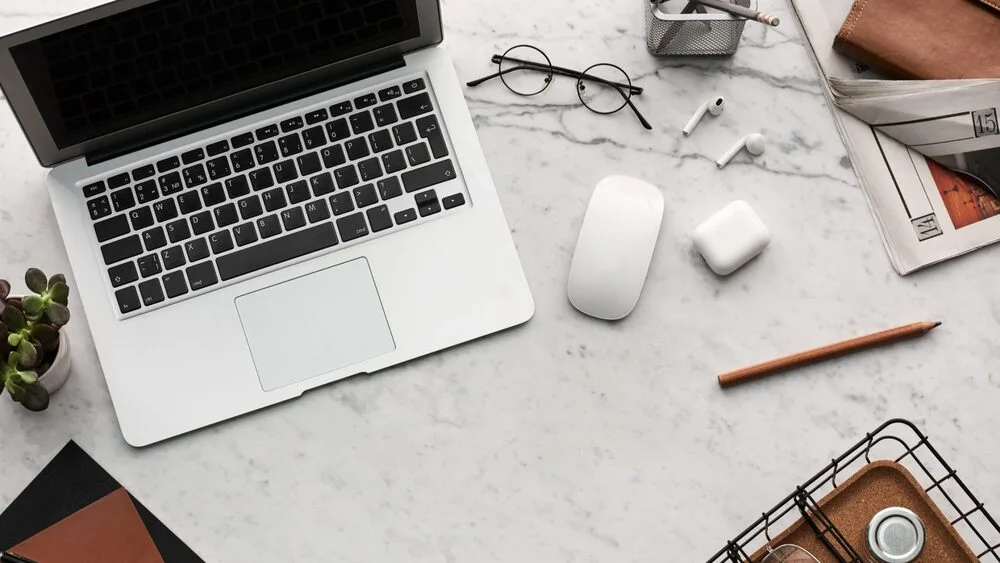 A marble desk with an open laptop, glasses, wireless earbuds, a mouse, a pencil, notebooks, and a small plant.