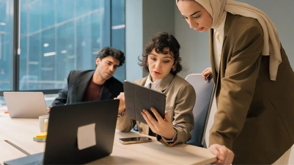 Three people in an office setting. Two women and a man are gathered around a table with laptops and a tablet. One woman shows the tablet to the others. Large windows in the background let in natural light.