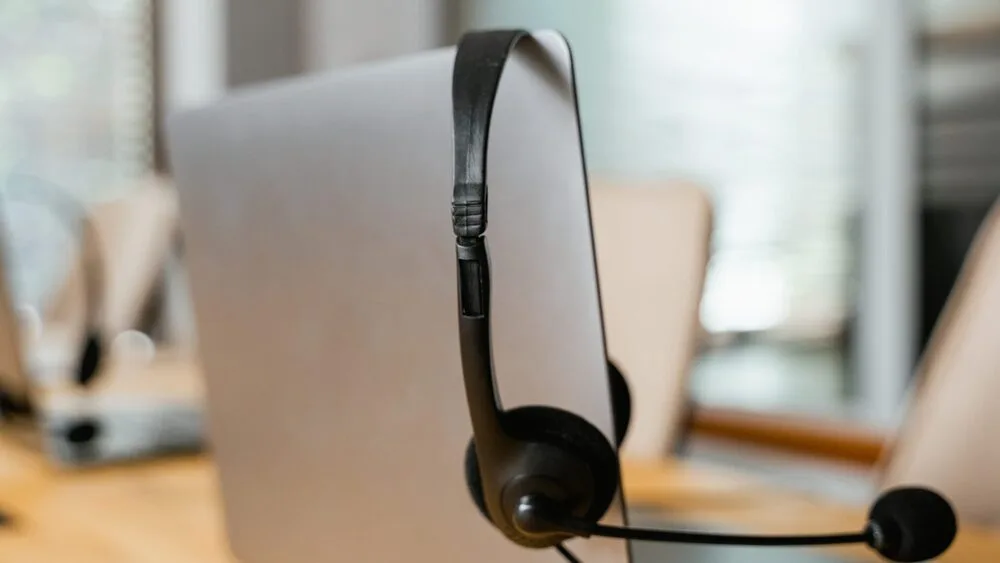 A black headset rests on the edge of a gray laptop in an office setting. The background features blurred office chairs and a conference table, suggesting a workplace or call center environment.