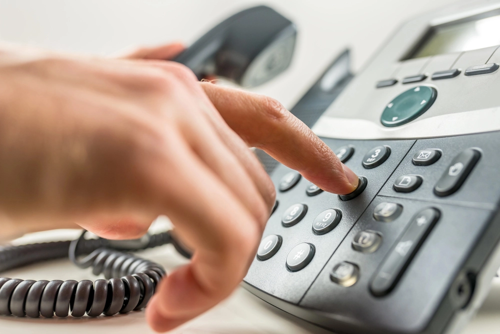 A close-up of a person's hand dialing a number on a landline telephone. The phone is off the hook, and the display and buttons are visible. The finger is pressing a button on the keypad.