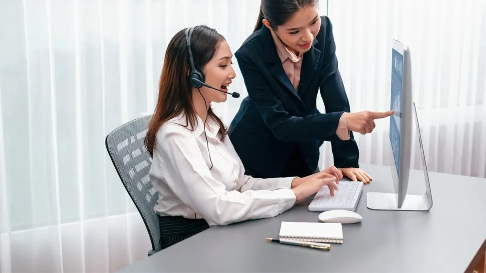 Two women in an office setting, one seated and wearing a headset, typing on a keyboard. The other stands beside her, pointing at the computer screen.