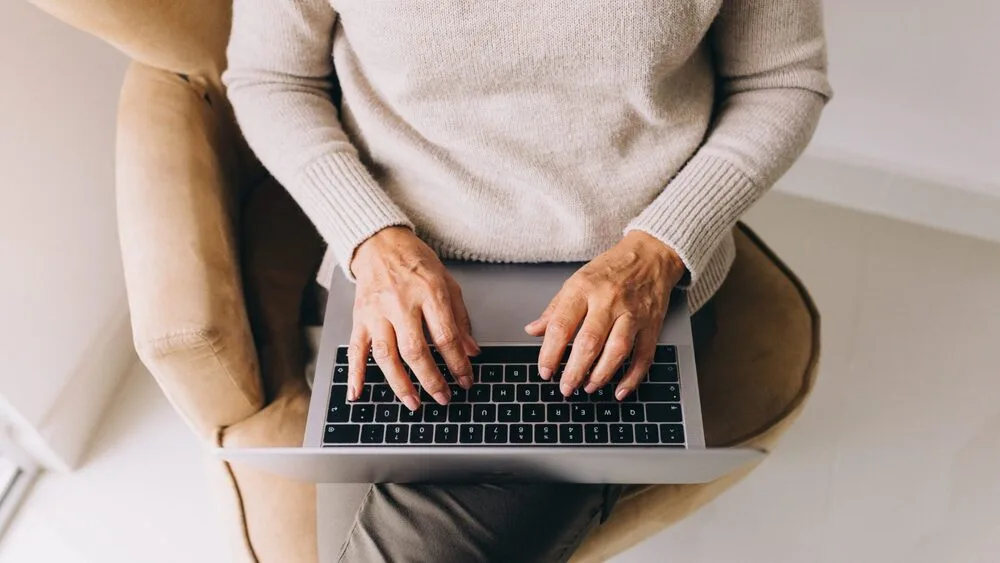 A person wearing a beige sweater and sitting on an armchair types on a laptop placed on their lap. 