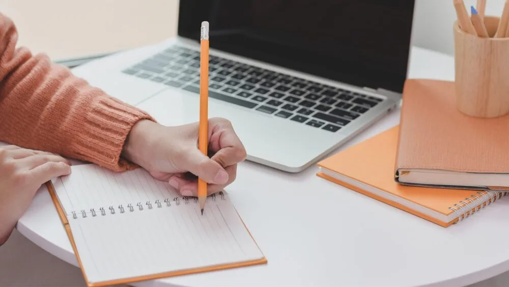 A person in an orange sweater writes in a lined notebook with a pencil. A laptop is open on a round white table.