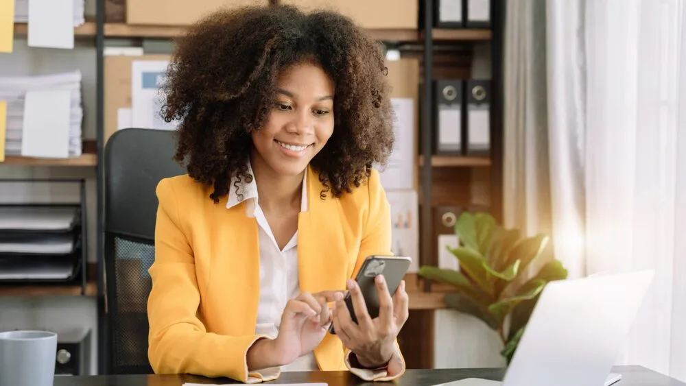 A person wearing a yellow blazer sits at a desk using a smartphone, with a laptop open in front of them. 