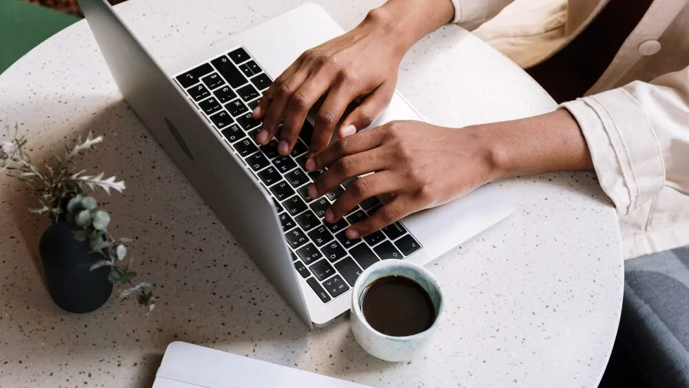 A person types on a laptop at a round, speckled table, with a cup of coffee nearby. 