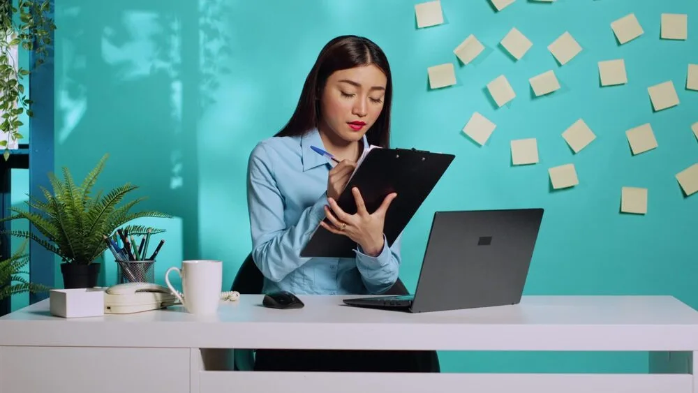 A person sits at a desk with a laptop, writing on a clipboard.