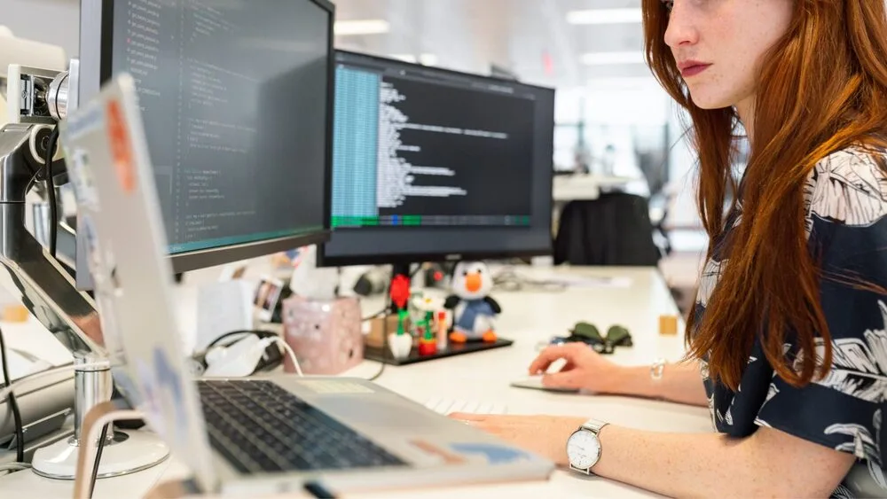 A person with long red hair is working at a white desk with a laptop and two monitors displaying code.