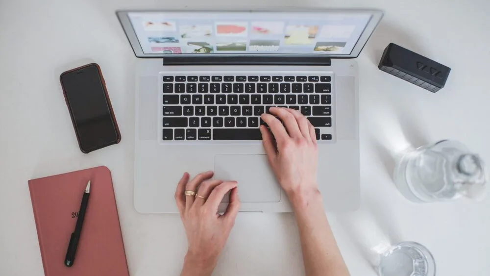 A person types on a laptop at a desk. Nearby are a smartphone, a closed notebook with a pen on top, a small black speaker, and a glass of water. 