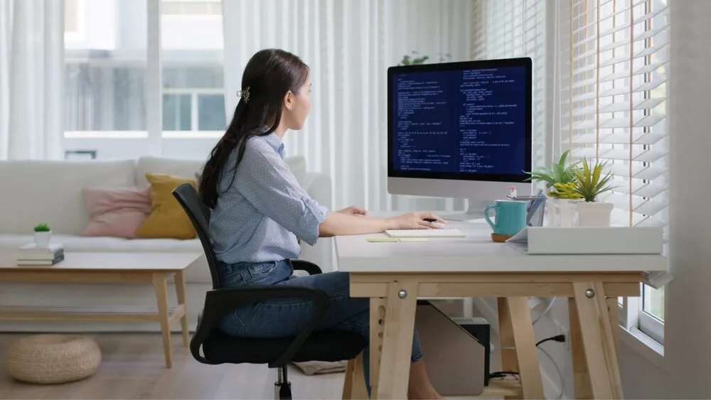 A person sits at a wooden desk in a bright room, typing code on a large computer monitor.