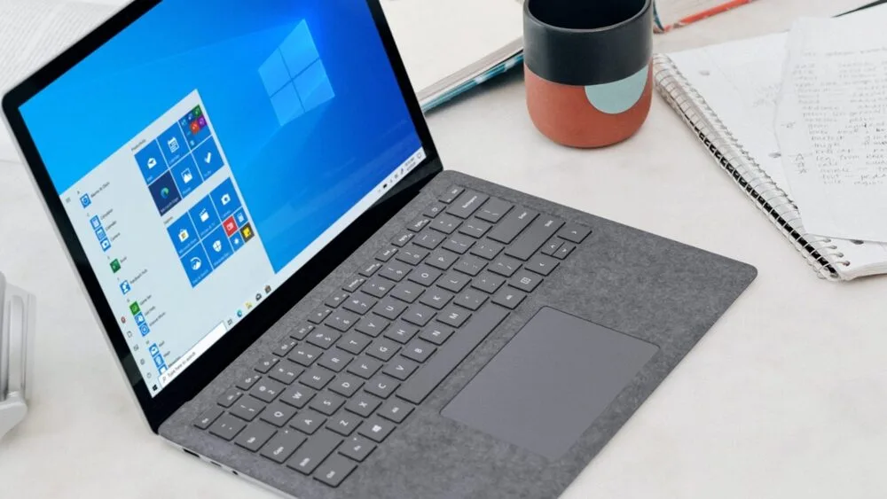 A laptop with a gray keyboard and touchpad is open on a white table displaying the Windows 10 home screen.