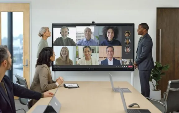 A group of four people in a conference room having a video call with five others displayed on a large screen.