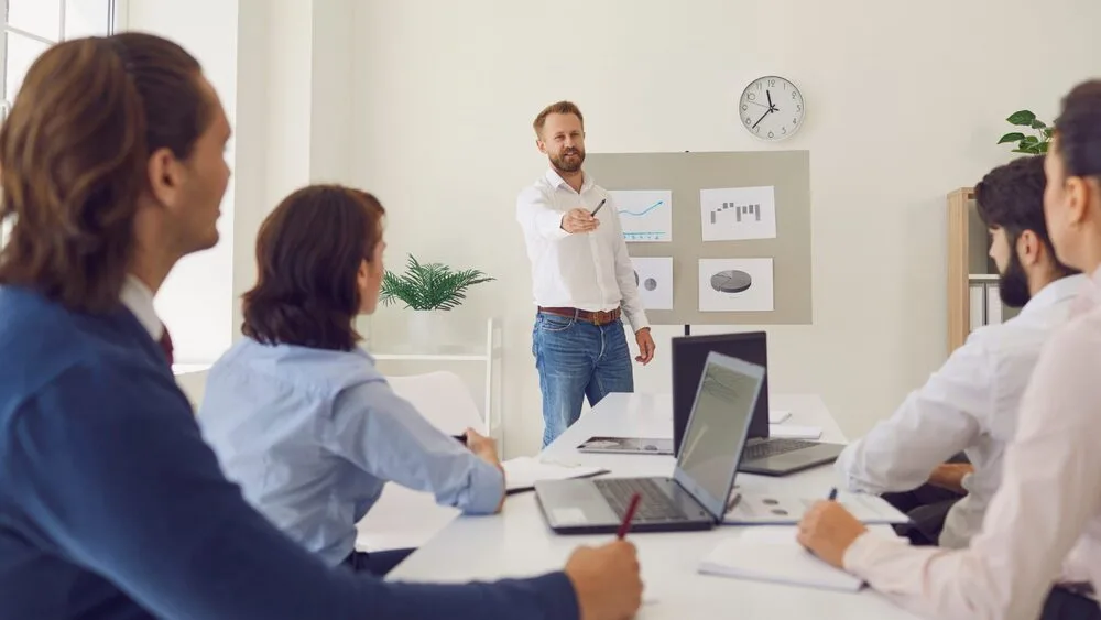 A man stands pointing at a flip chart during a meeting in a conference room.