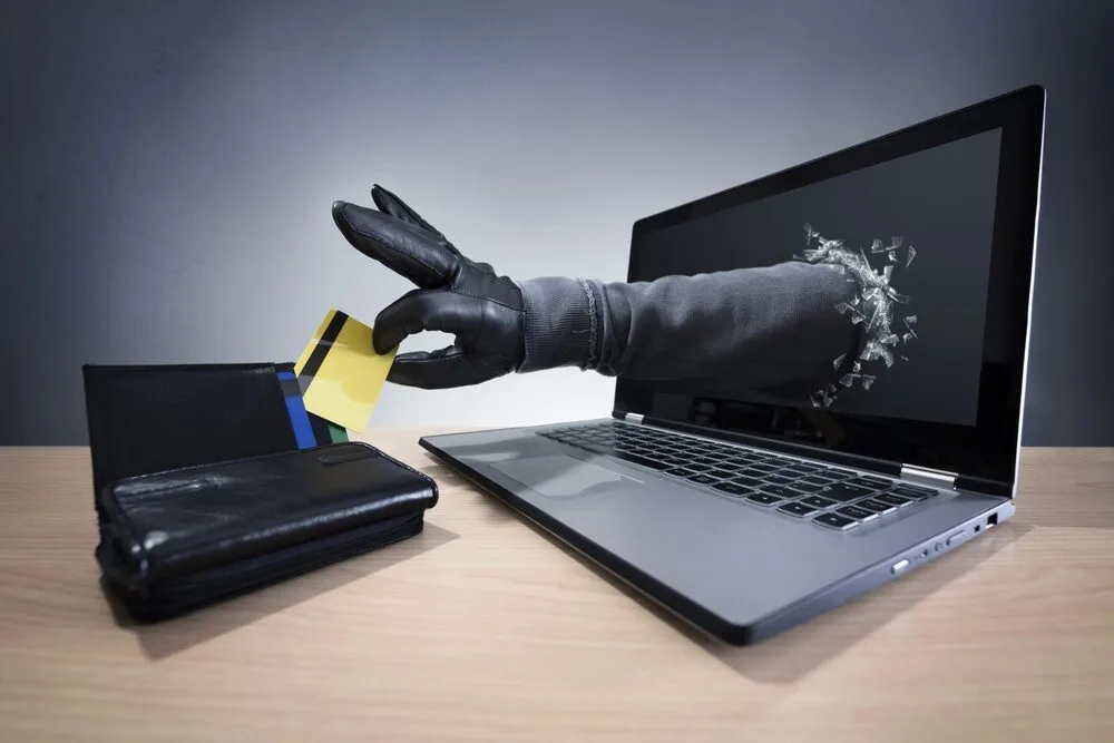 A gloved hand emerges from a laptop screen, reaching for a credit card next to a wallet on a wooden table. 